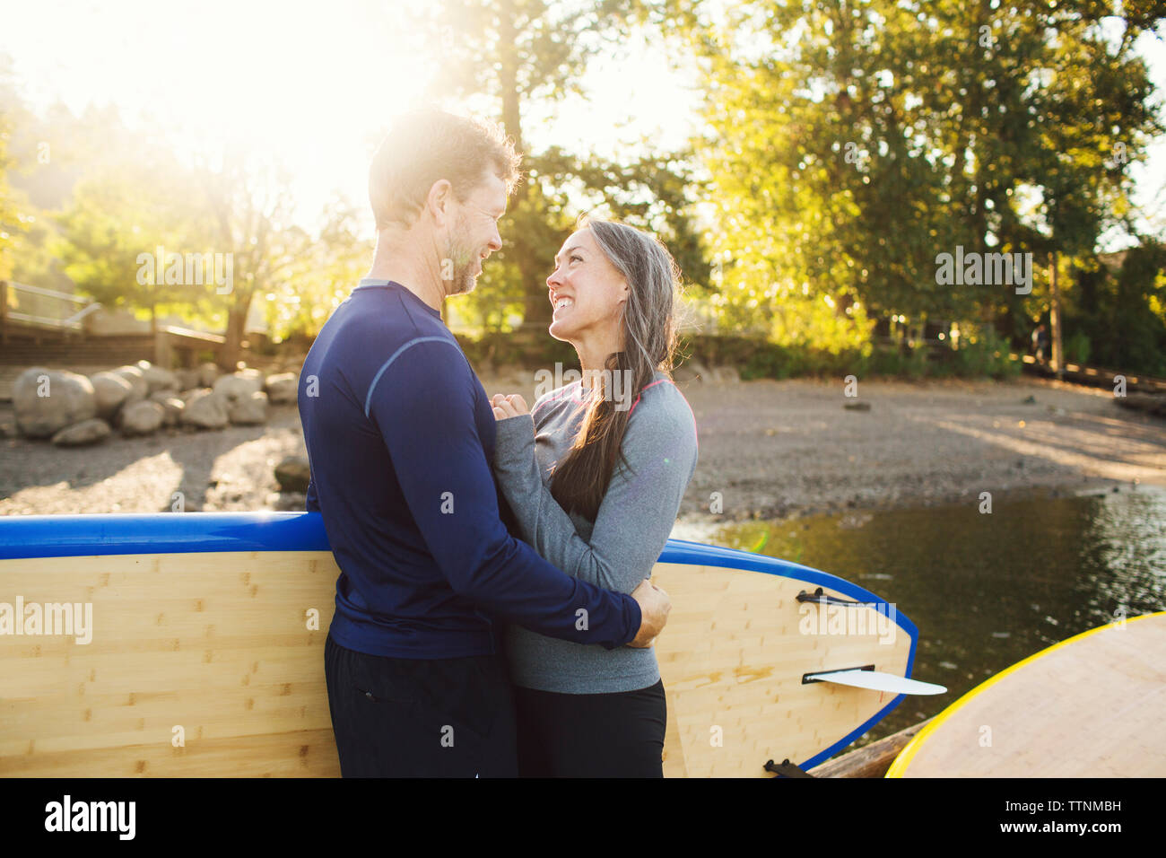 Smiling couple looking at each other while standing with paddleboards at riverbank Stock Photo