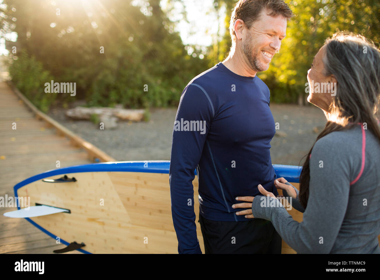 Cheerful couple with paddleboard looking at each other while standing on boardwalk Stock Photo
