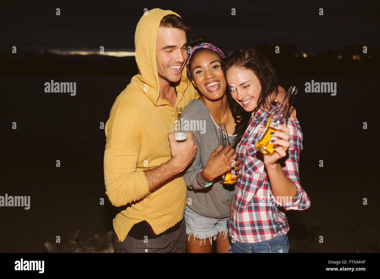 Smiling friends with arm around holding bottles while standing by river at night Stock Photo