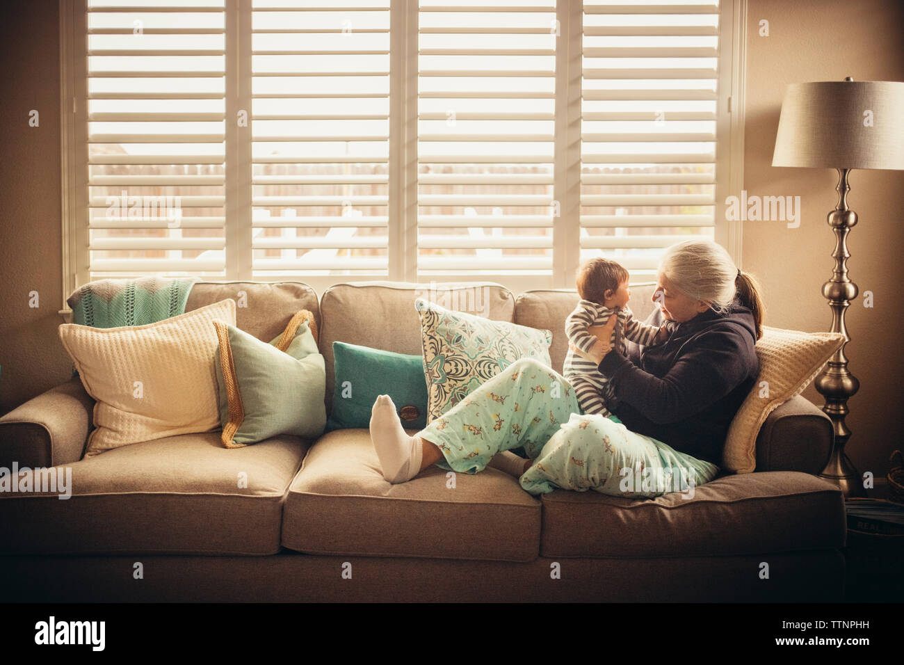 Grandmother playing with baby grandson on sofa against window Stock Photo