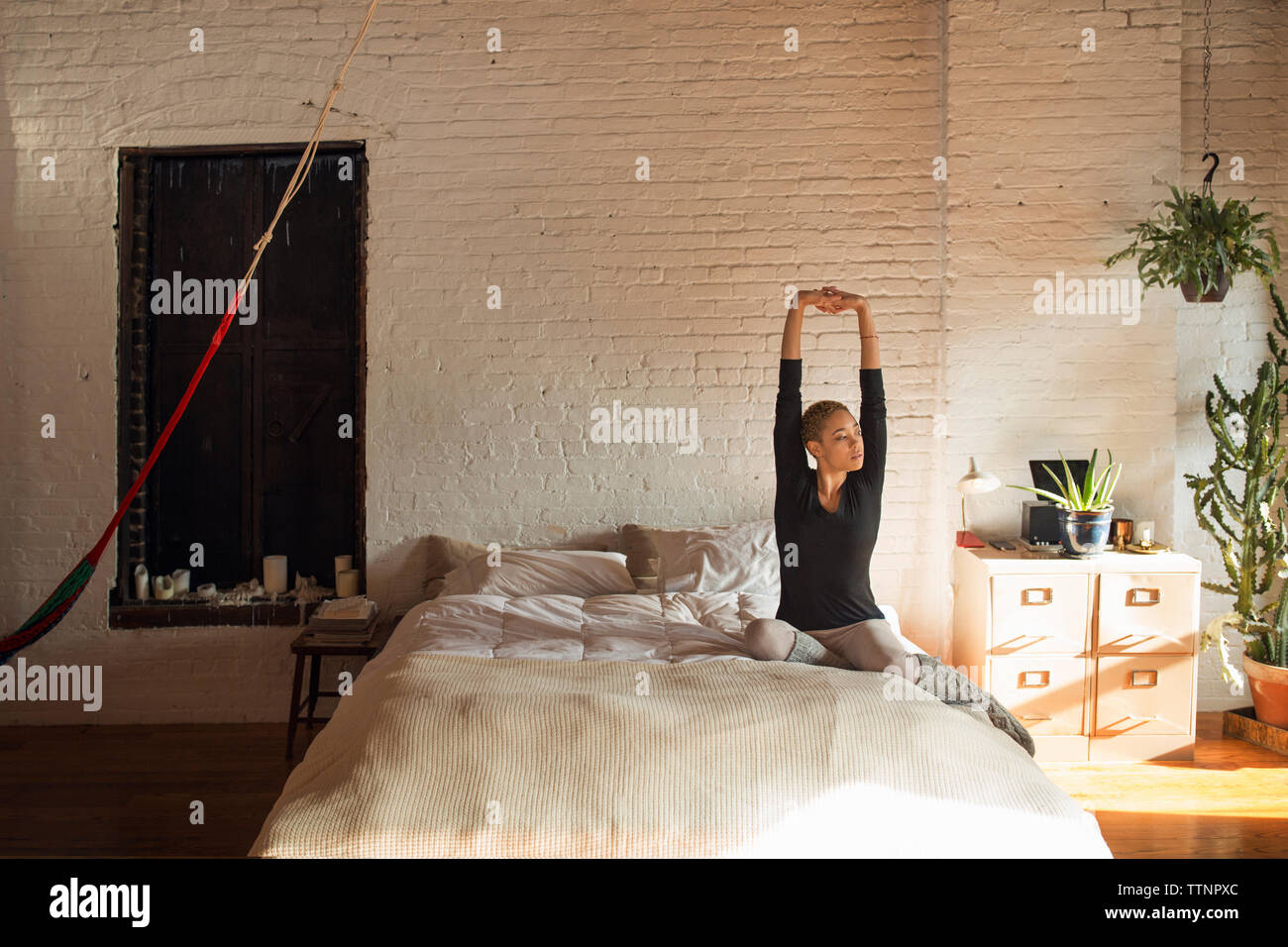 Young woman stretching arms while sitting on bed at home Stock Photo