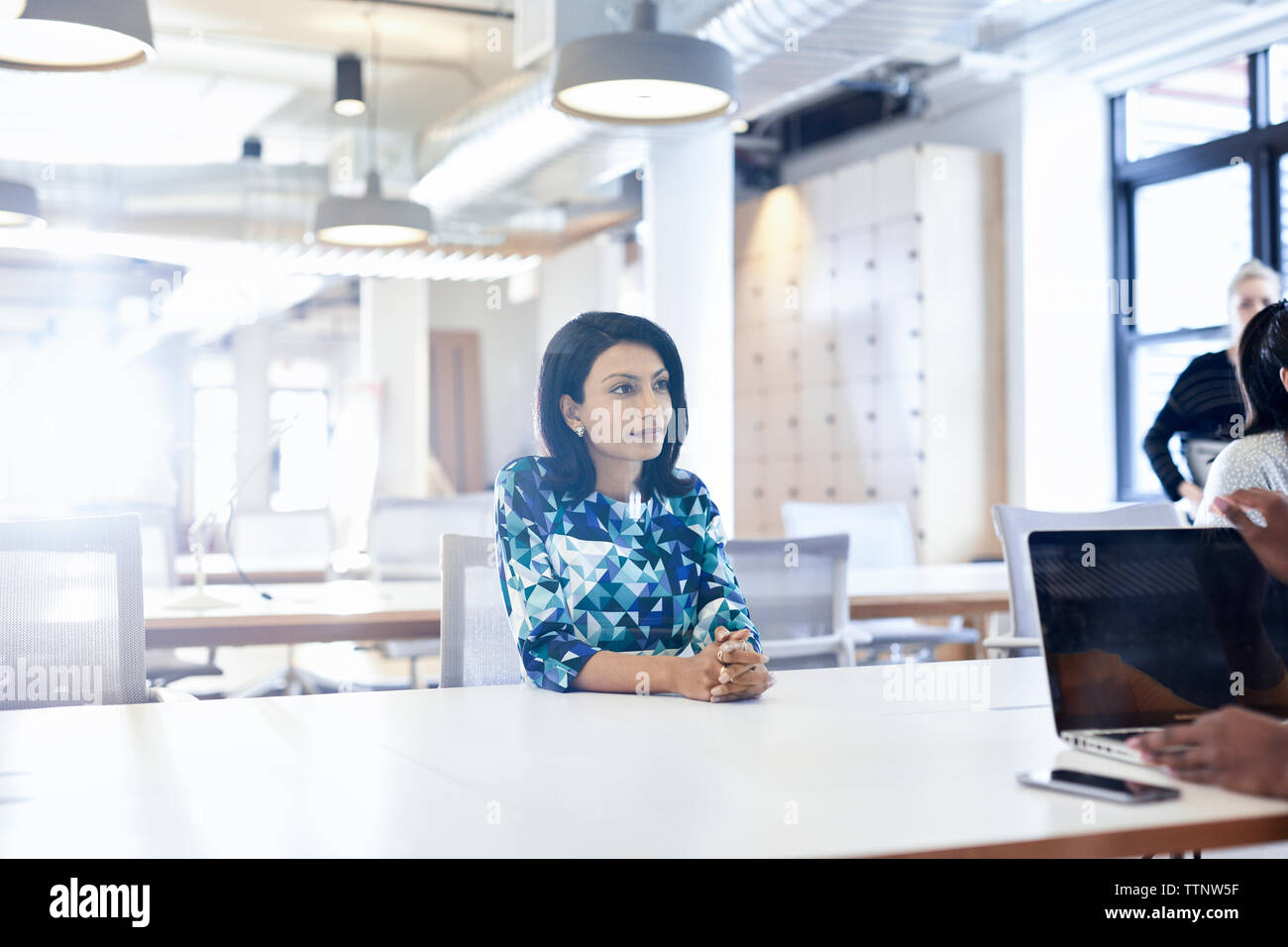 businesswoman with hands clasped looking away while sitting at conference table in creative office Stock Photo