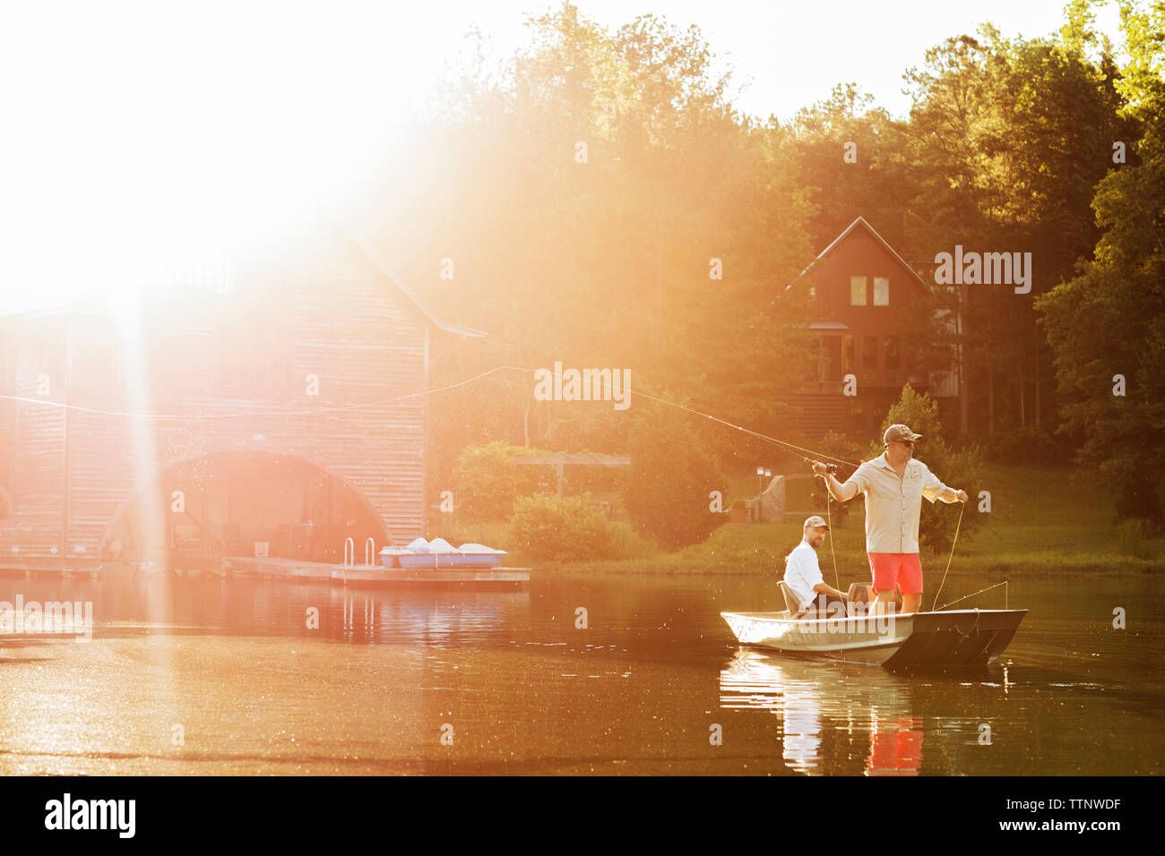 Man casting fishing line in lake with friend sitting in rowboat during sunny day Stock Photo