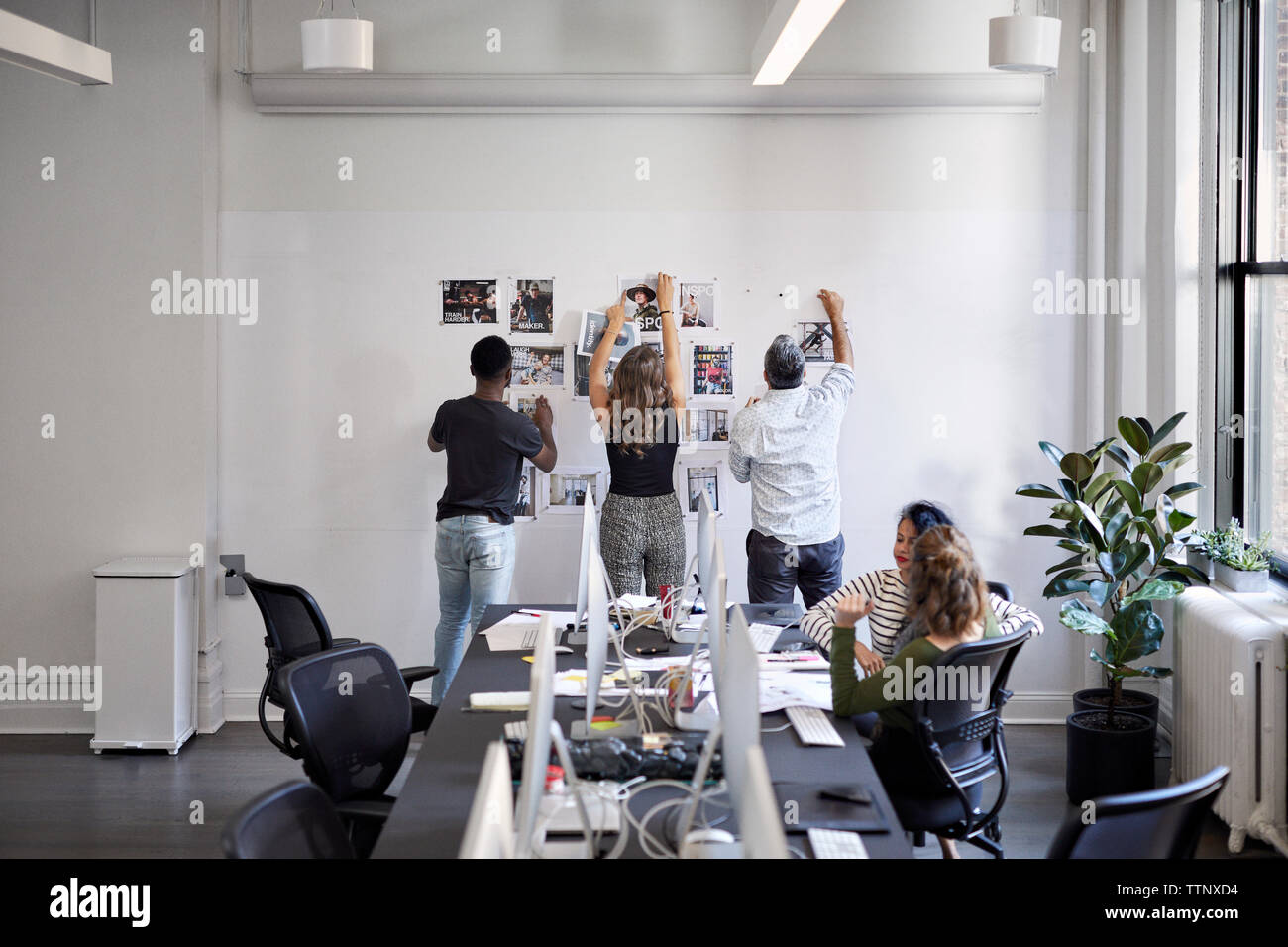 business people attaching photograph printouts on bulletin board while female colleagues sitting at conference table in office Stock Photo