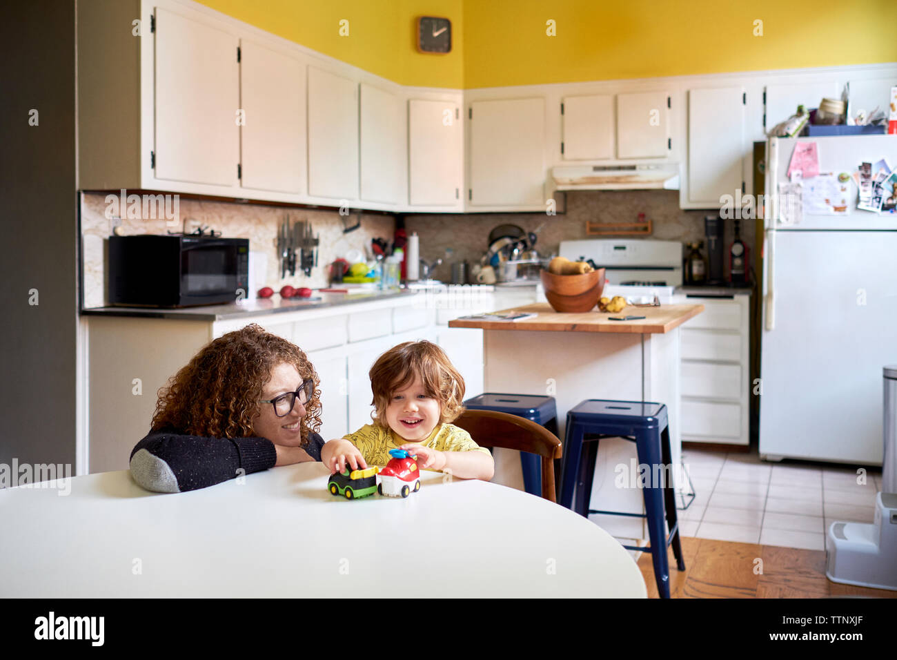 Mother looking at son playing with toys while sitting in kitchen Stock Photo