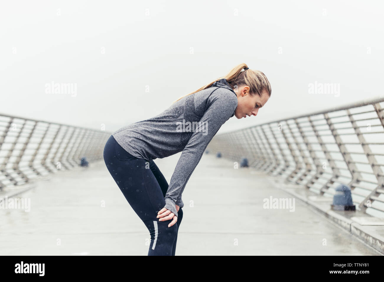 Side view of tired female athlete standing with hands on knees on bridge against clear sky Stock Photo