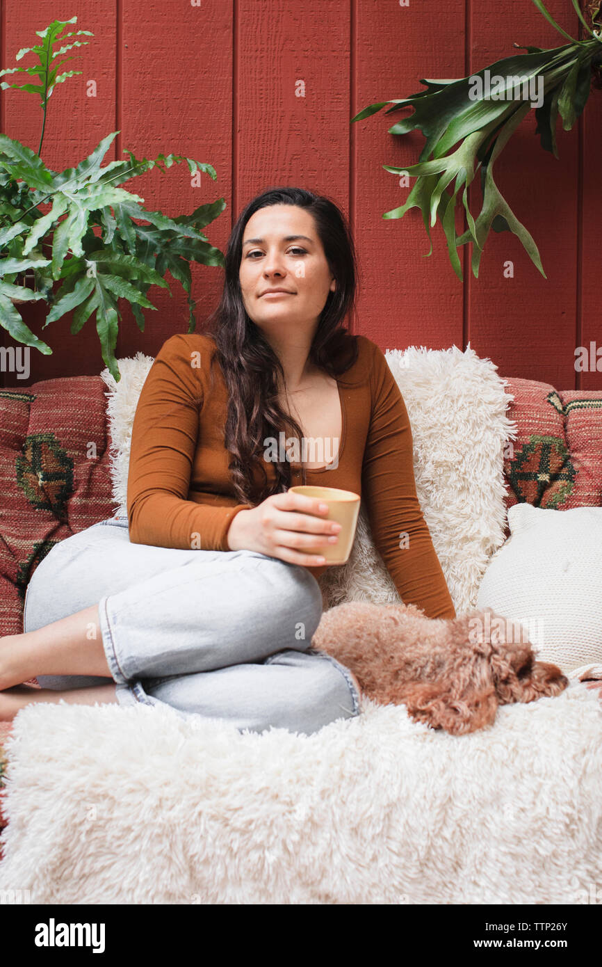 Portrait of confident young woman with poodle and drink resting on sofa by plants at porch Stock Photo