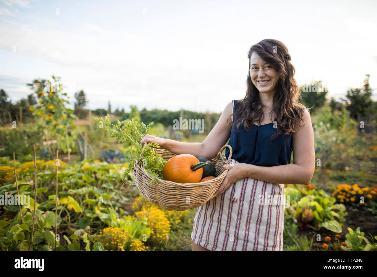 Portrait of cheerful woman carrying vegetables in basket against clear sky at community garden Stock Photo