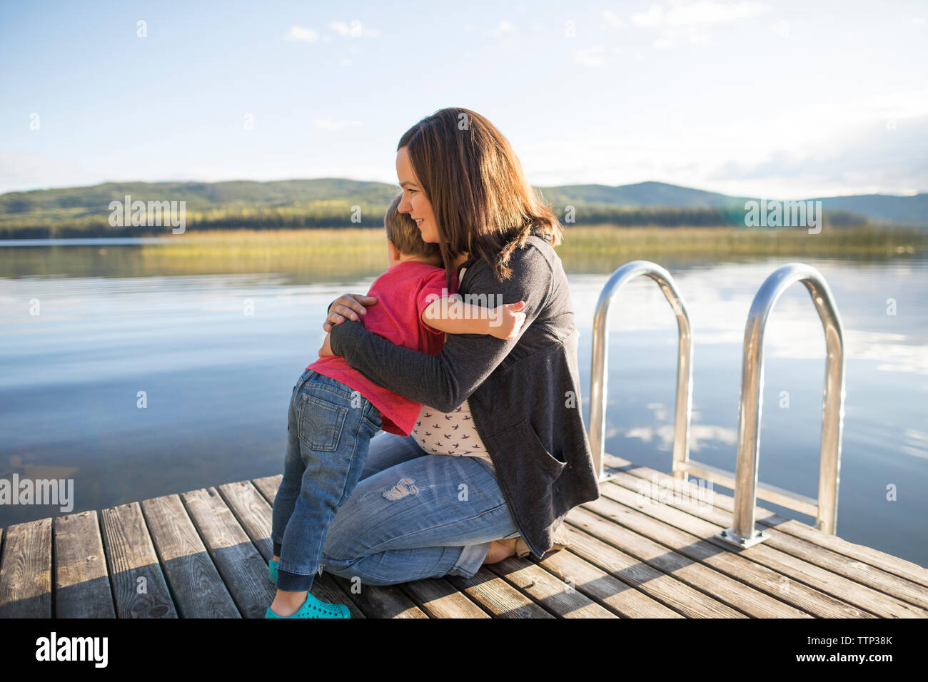 Loving son embracing pregnant mother on wooden pier over lake Stock Photo