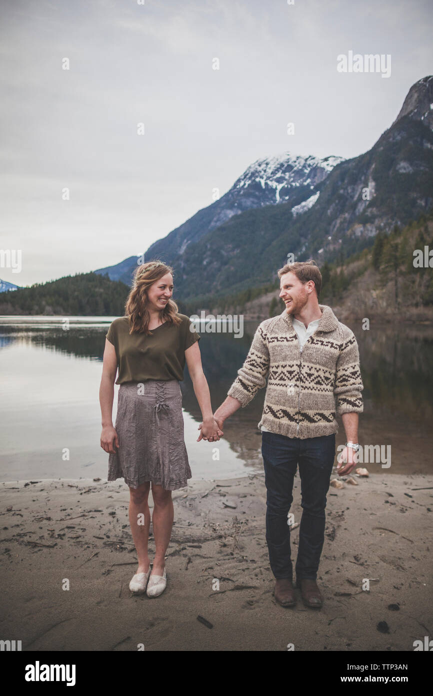 Full length of smiling young couple holding hands while standing on lakeshore at Silver Lake Provincial Park Stock Photo