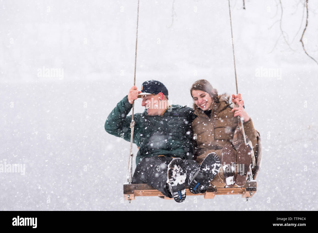 Couple laughing on swing over river during snowfall Stock Photo