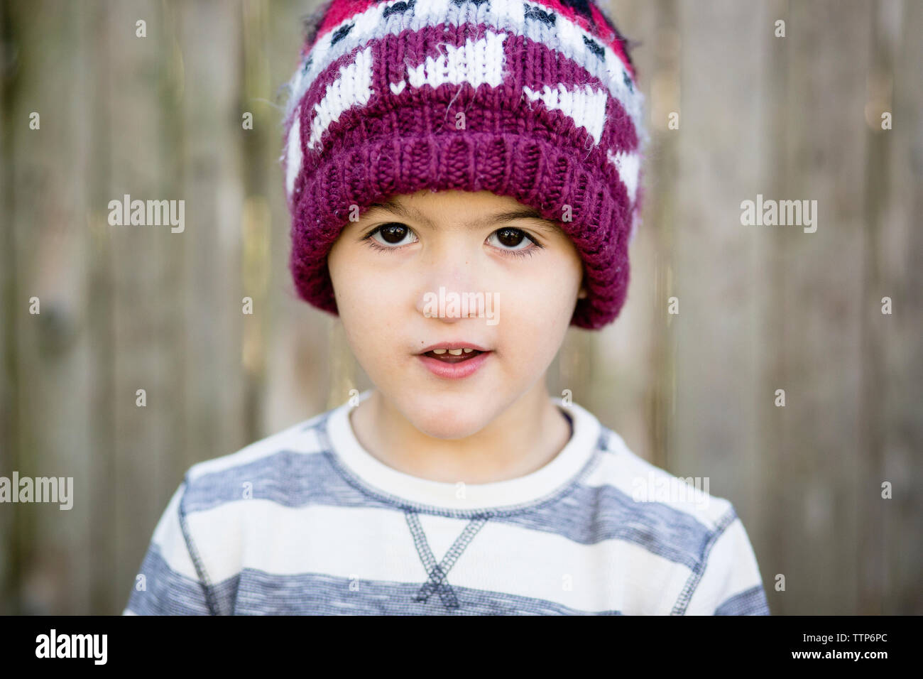 Close-up portrait of cute boy wearing knit hat while standing against fence in yard Stock Photo