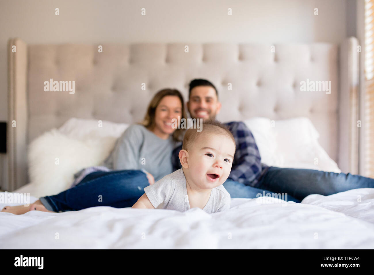 Baby boy laying on bed smiling with mom and dad in background indoors Stock Photo