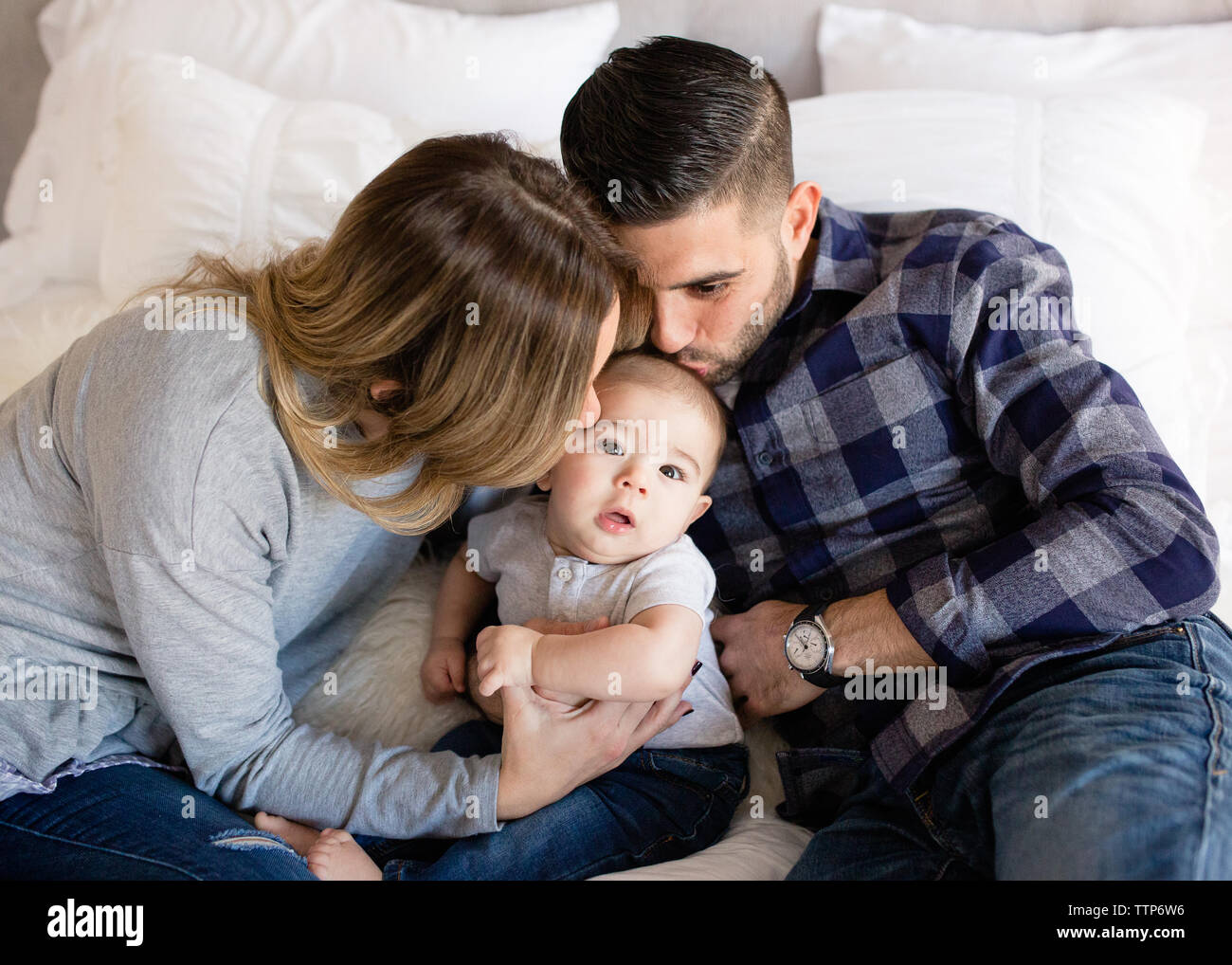 Mom and dad kissing cute baby boy on cheek indoors on bed Stock Photo