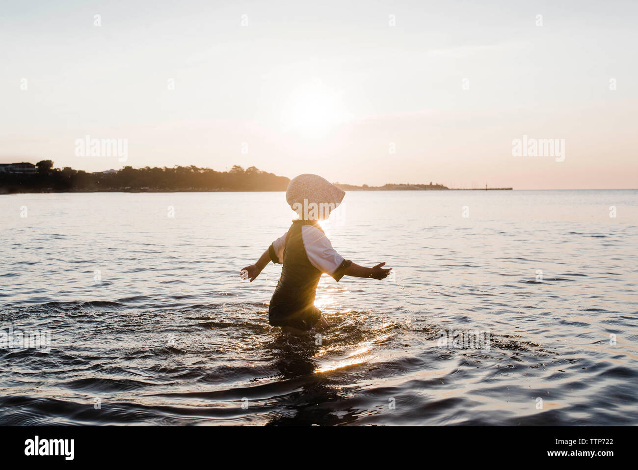 Side view of girl wearing cap while walking in sea against sky during sunset Stock Photo