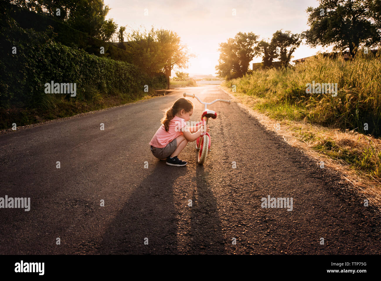Little girl repairs balance bike in road at sunset with backlight Stock Photo
