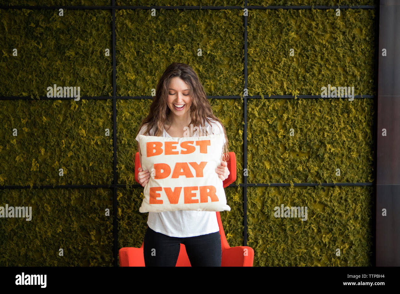 Happy young woman holding cushion with best day ever text while standing against wall at home Stock Photo