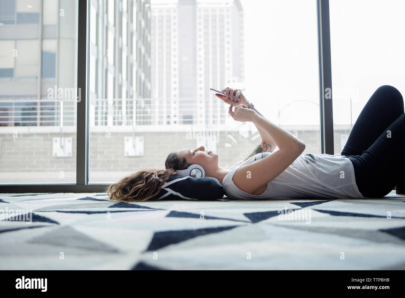 Young female using smart phone while lying on carpet by window at home Stock Photo