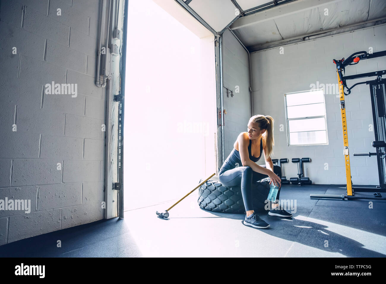 Thoughtful woman sitting on tire while holding water bottle in gym Stock Photo