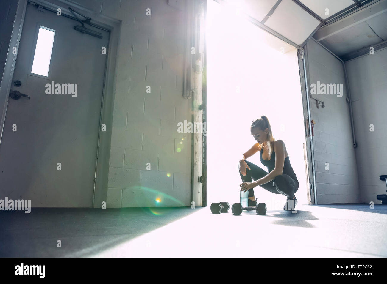 Tired woman holding water bottle while crouching in brightly lit gym ...