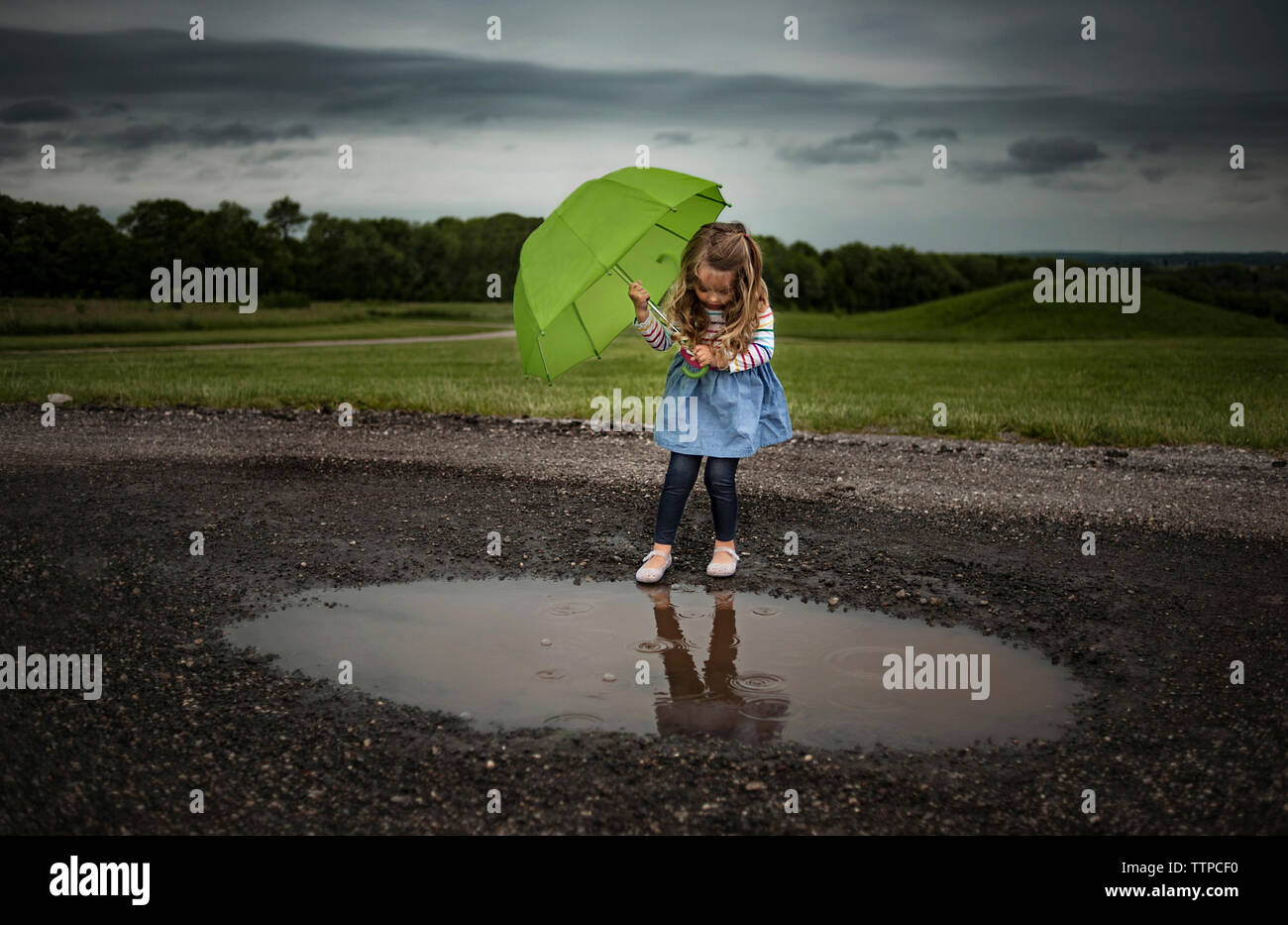 Girl holding umbrella while walking into puddle on road against stormy clouds Stock Photo