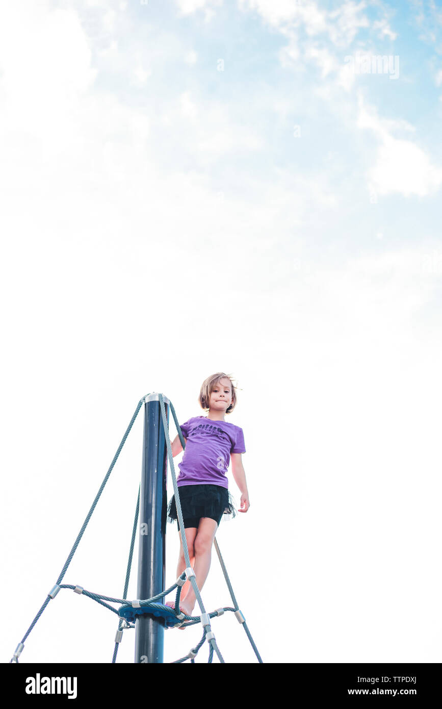 Low angle view of girl standing on outdoor play equipment against cloudy sky at playground Stock Photo