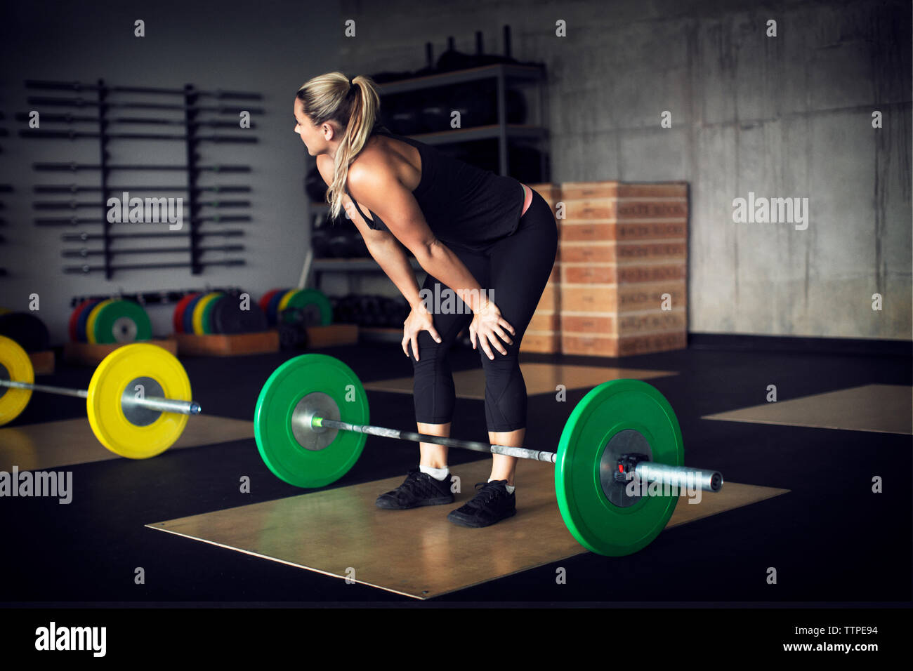 Tired female athlete standing with hands on knees by barbell in gym Stock Photo