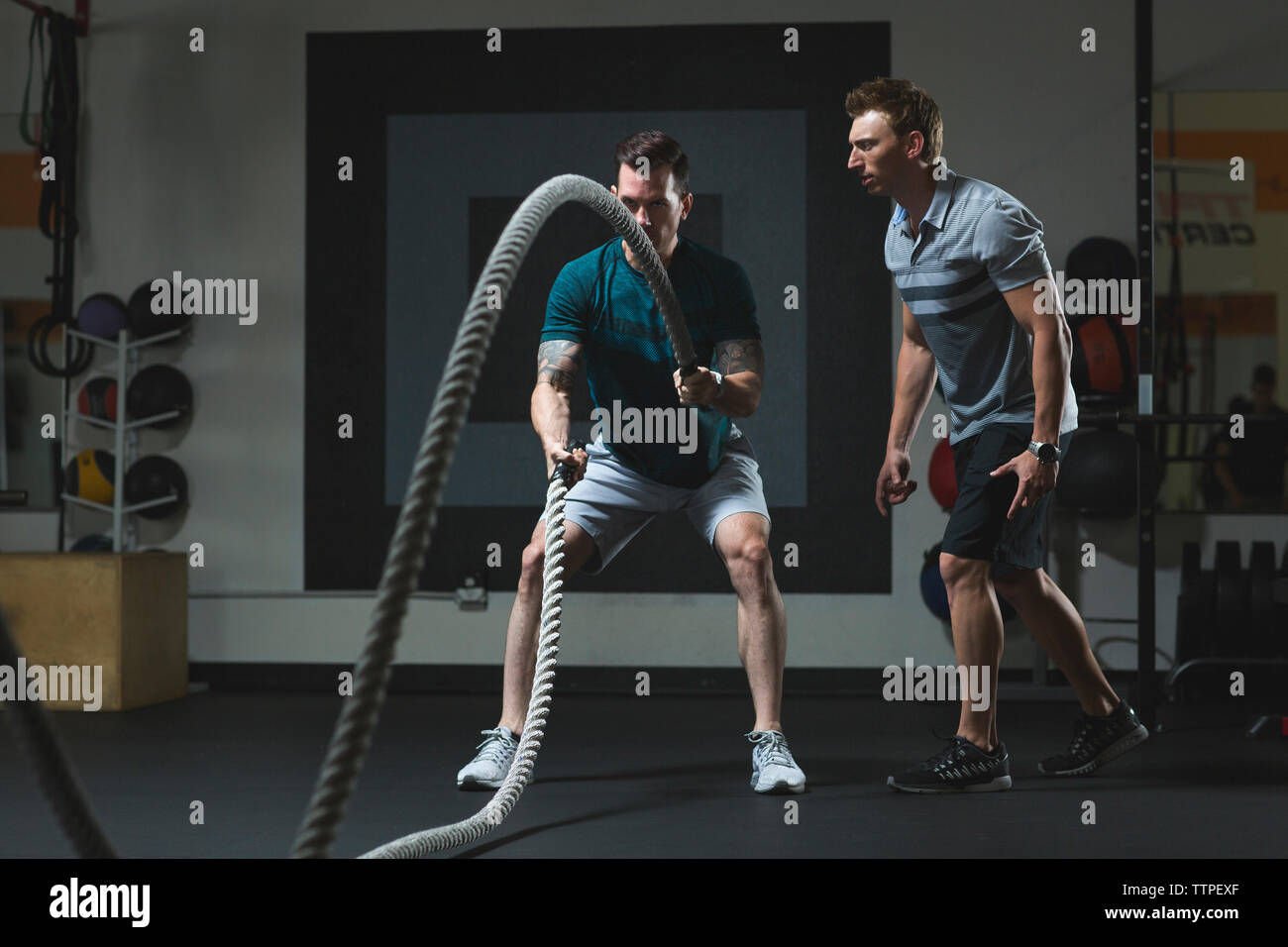 Trainer looking at male customer exercising with ropes in gym Stock Photo