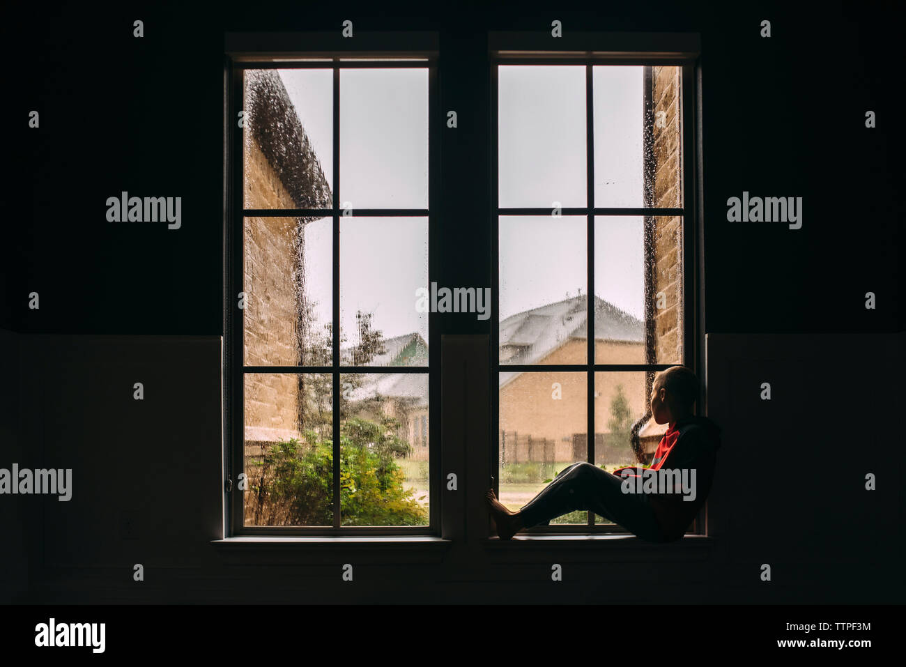 Side view of boy looking through window while sitting at home during rainfall Stock Photo