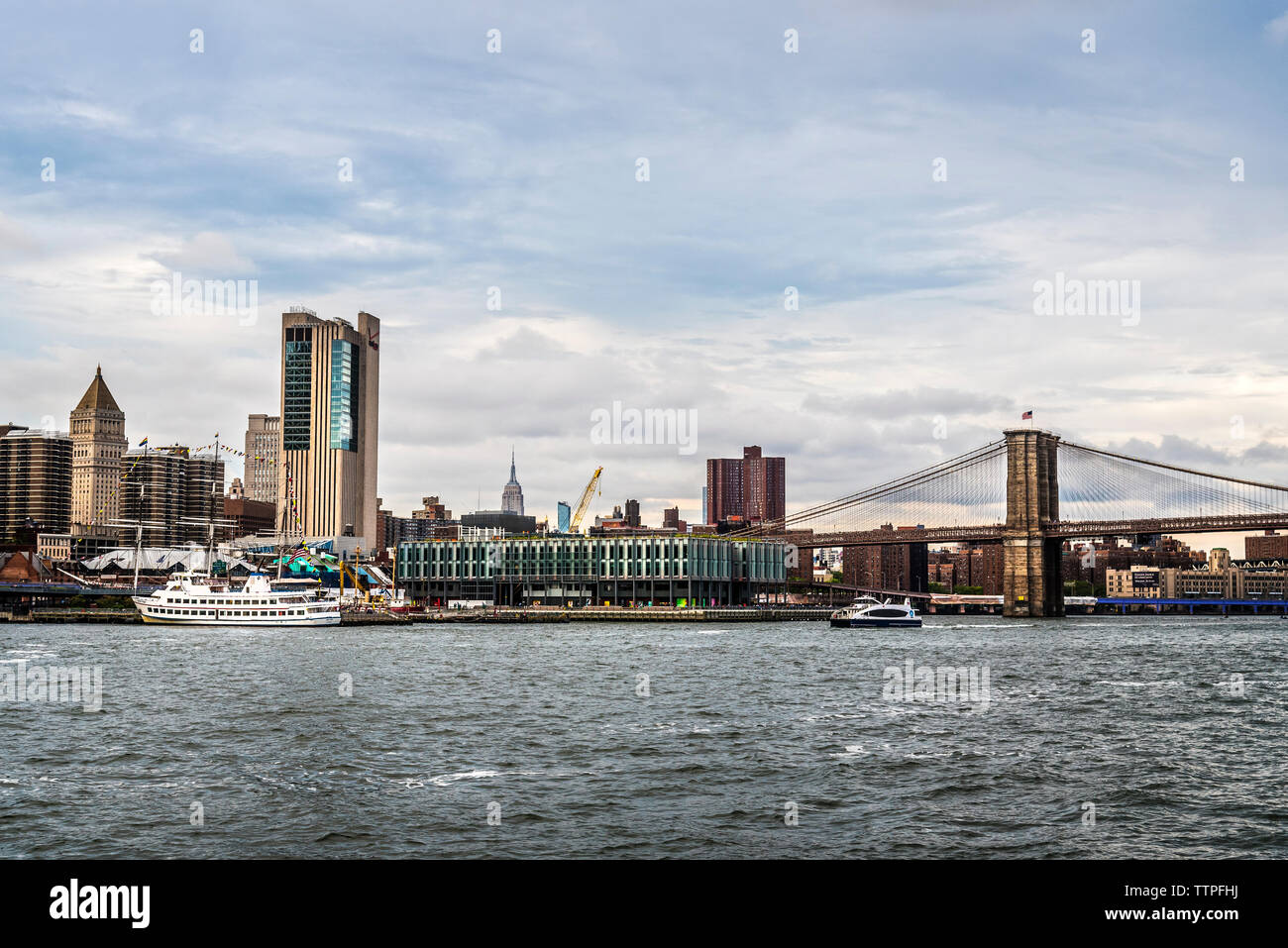 Brooklyn Bridge over East River by city against cloudy sky Stock Photo