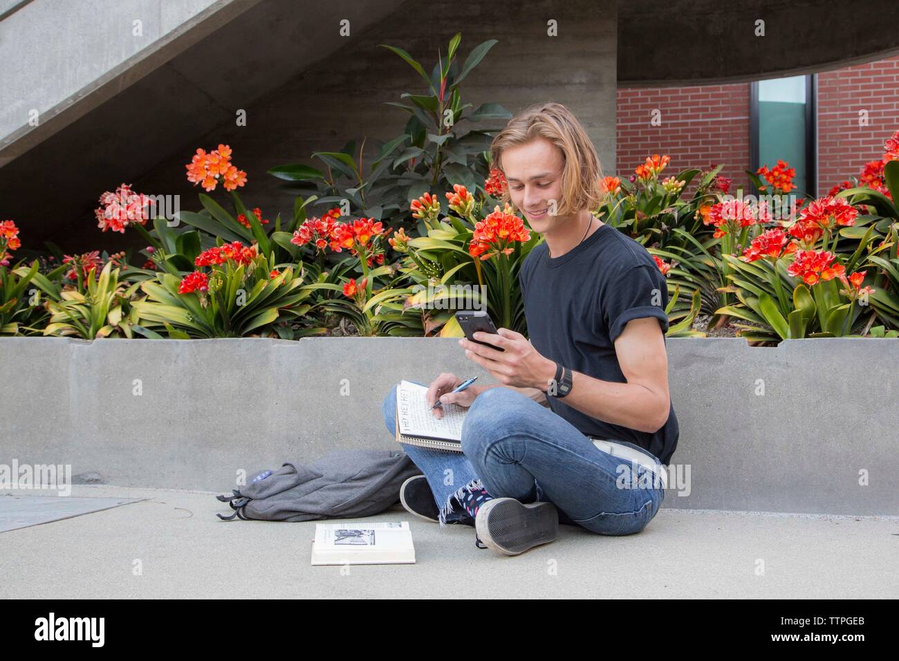 Male student sitting by flowers with books, smiling at text message Stock Photo