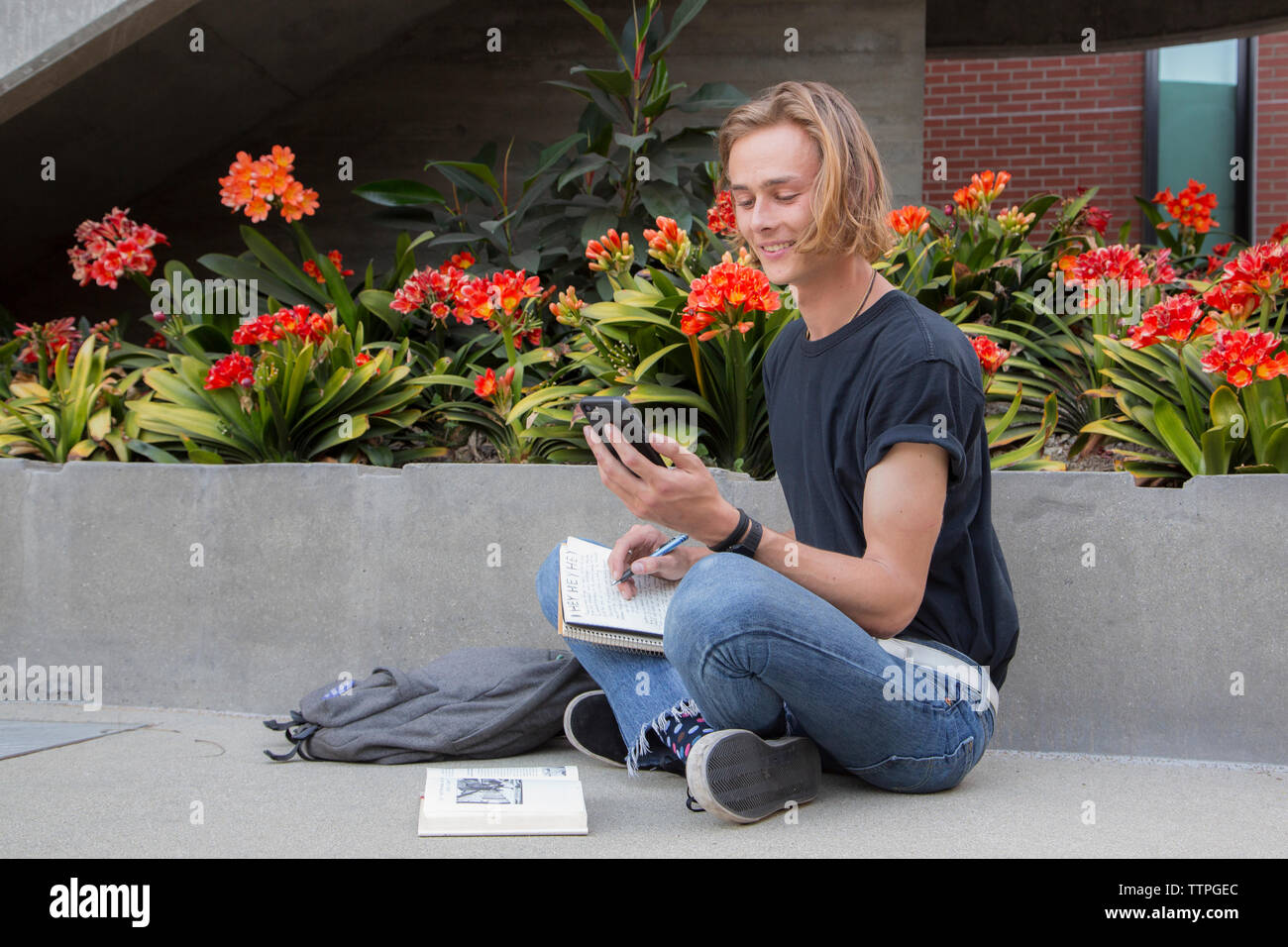 Male student sitting down with books and smiling at text message Stock Photo