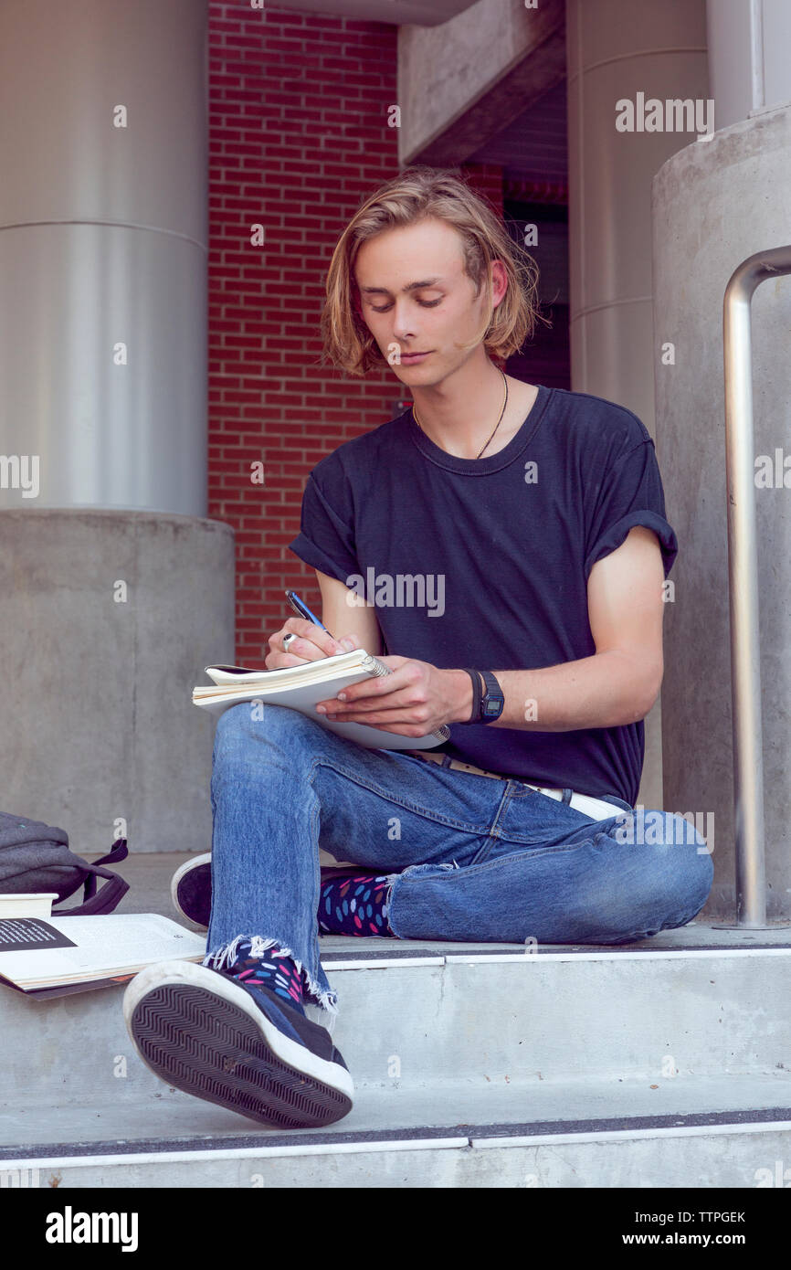 Male student sitting on stairs and writing on notebook Stock Photo