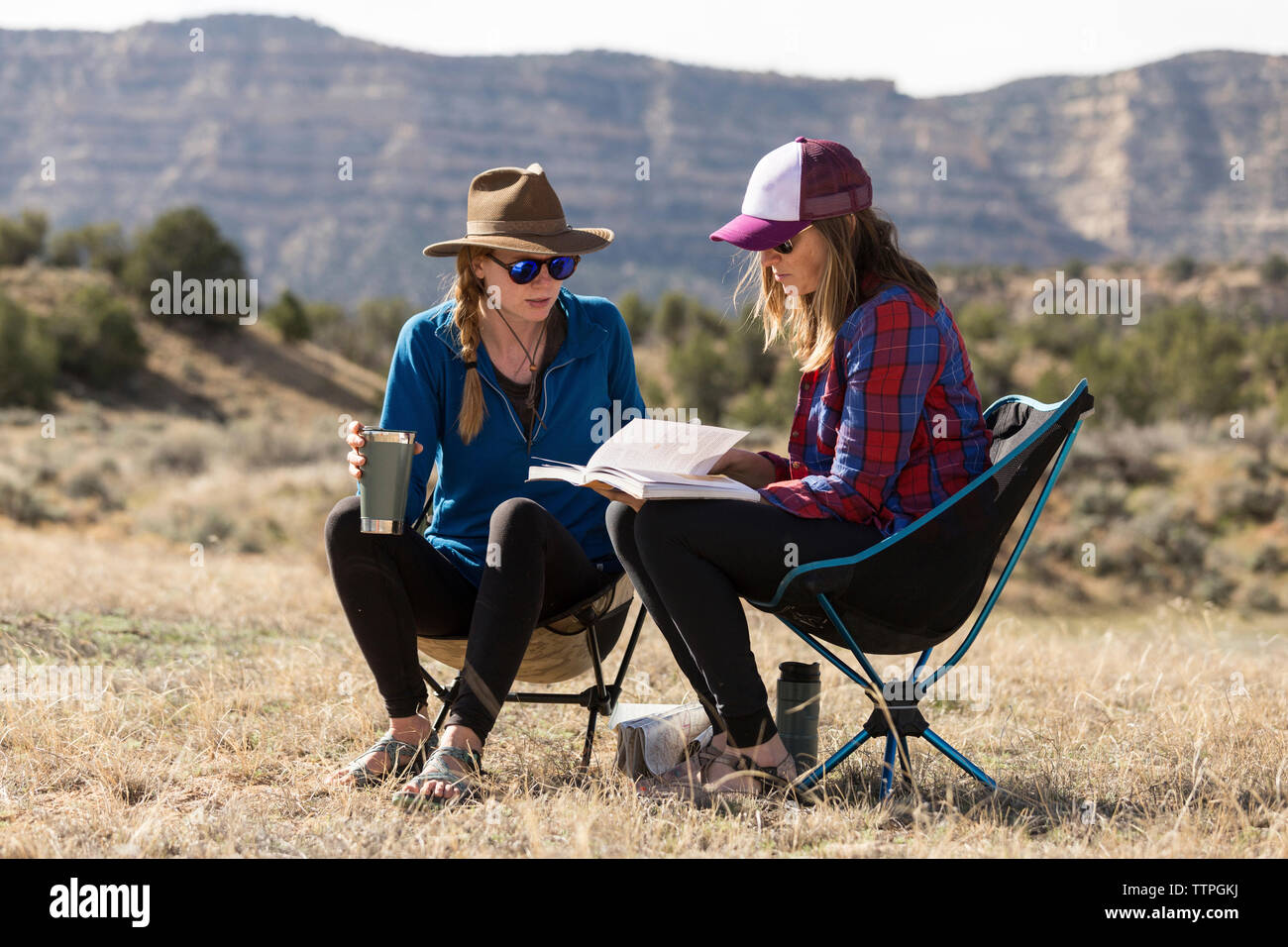 Female friends looking at book while sitting on camping chairs at field against mountains during sunny day Stock Photo