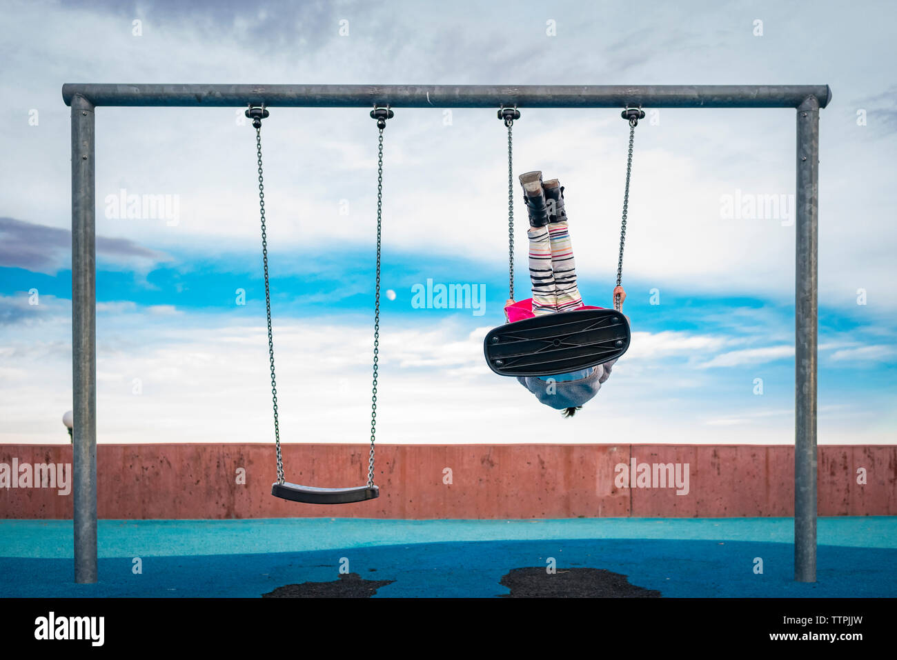 Playful girl swinging against cloudy sky at playground Stock Photo