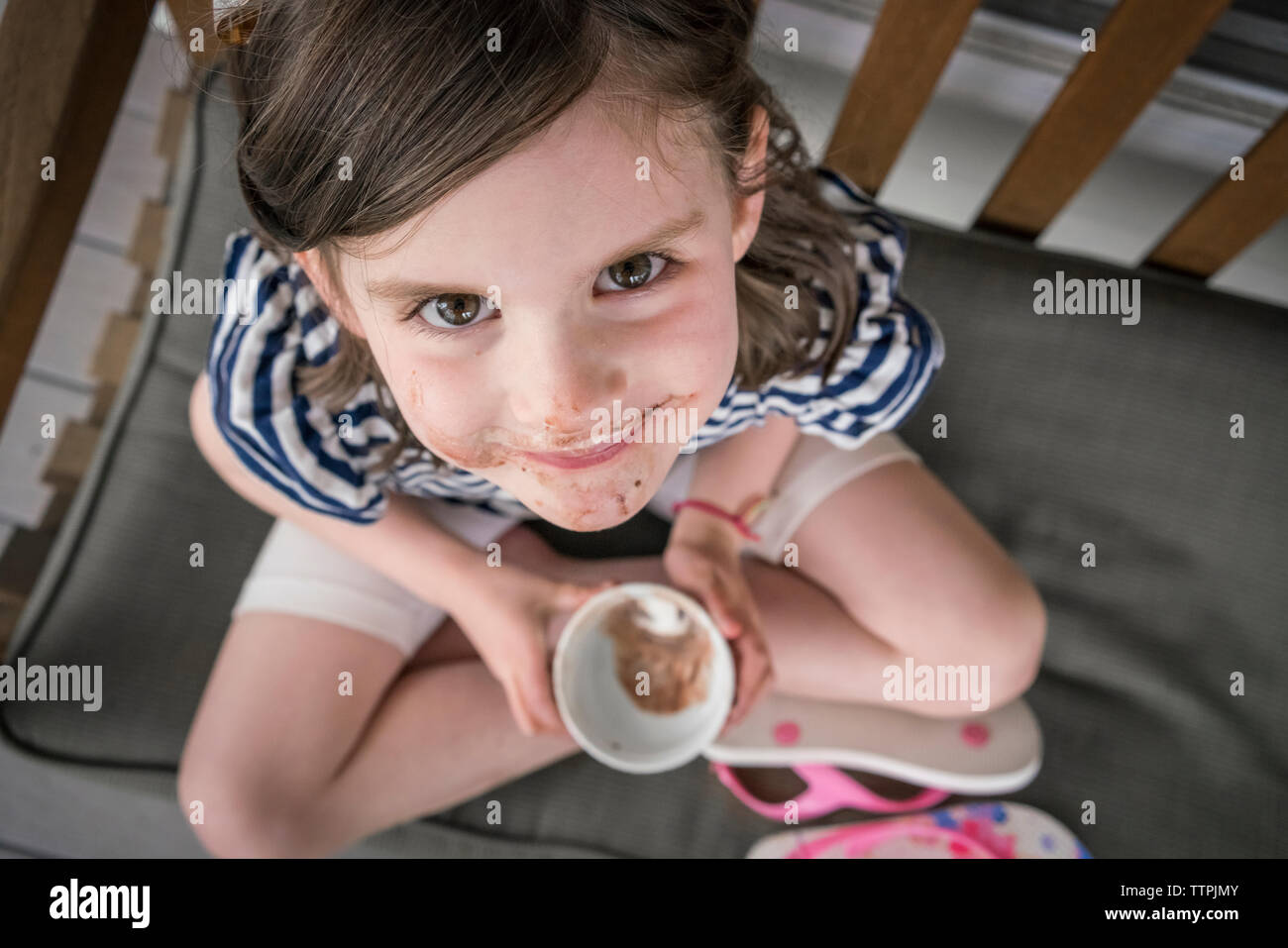 High angle portrait of cute girl with messy mouth eating chocolate while sitting on chair at home Stock Photo