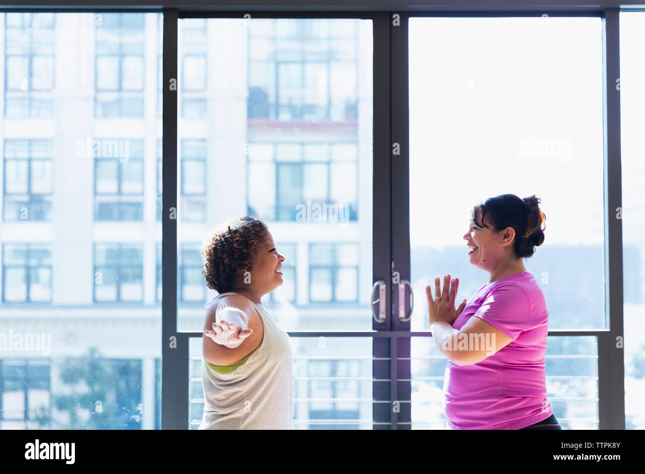 Happy friends exercising face to face while standing against window in yoga studio Stock Photo