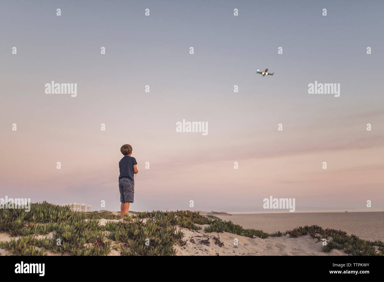 Rear view of boy looking quadcopter while standing at beach against sky Stock Photo