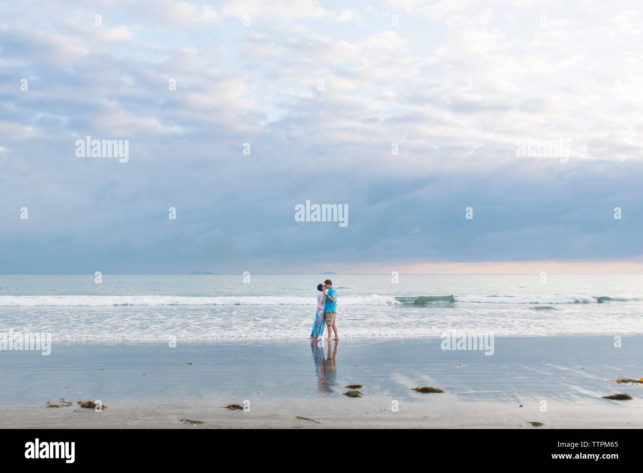 Loving couple standing on shore at beach against cloudy sky Stock Photo