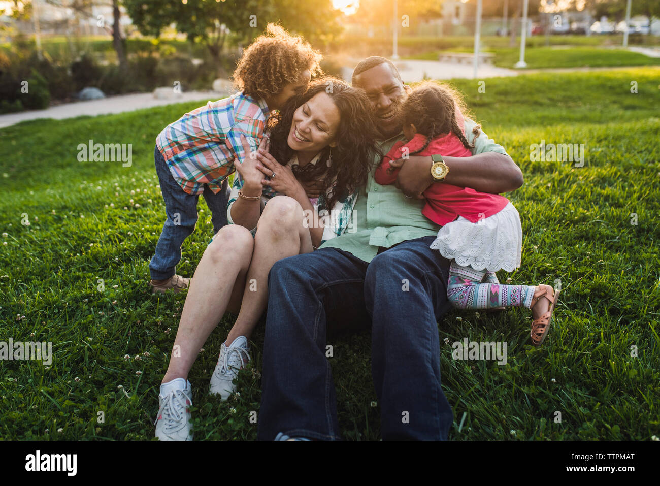 Children embracing parents on grassy field at park during sunset Stock Photo