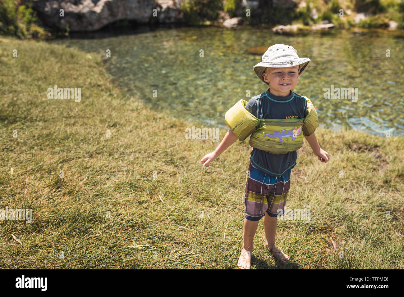Full length of cute smiling boy with water wings standing on lakeshore during sunny day Stock Photo