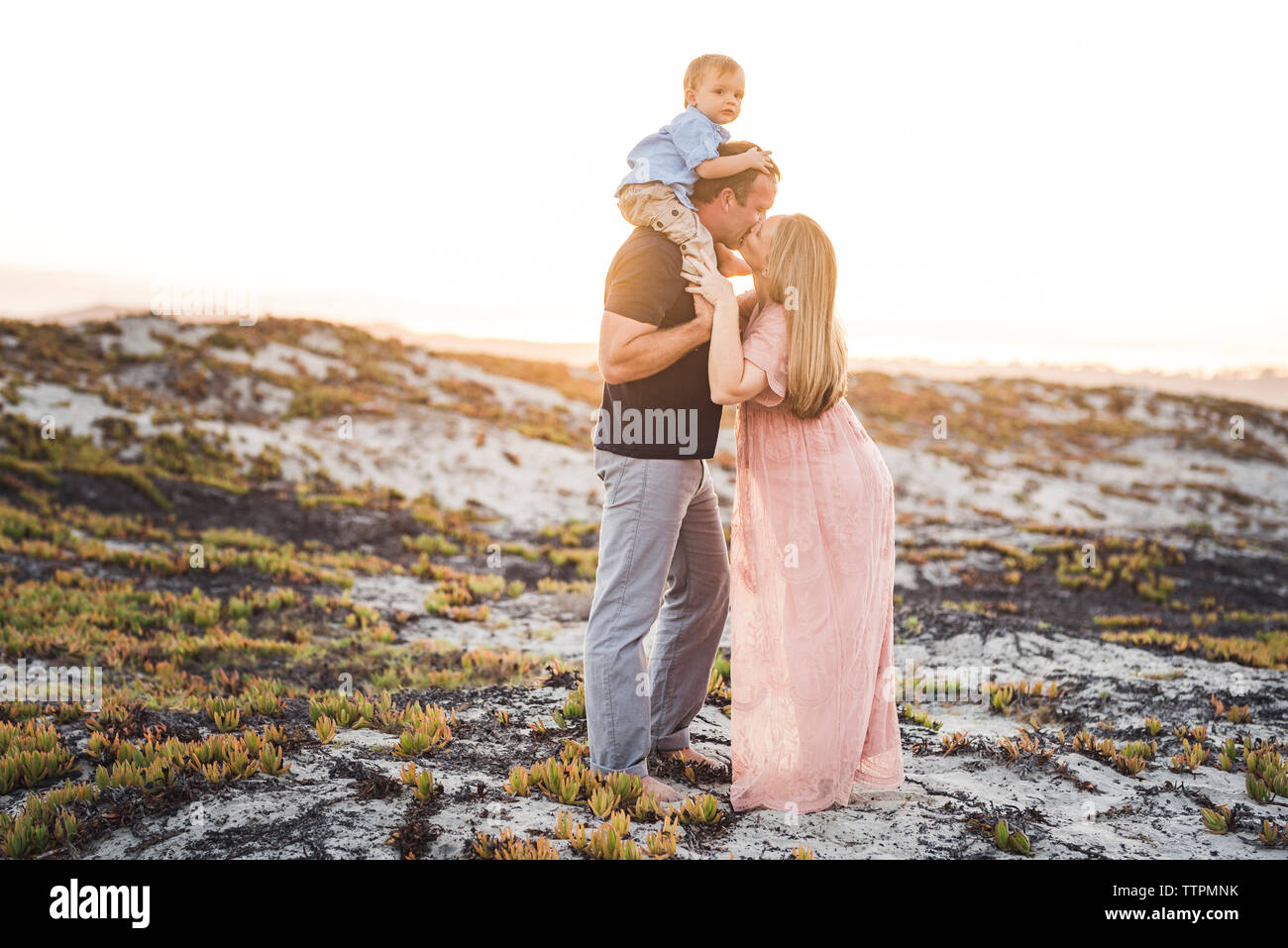 Husband kissing wife while carrying son on shoulders at beach against clear sky during sunset Stock Photo