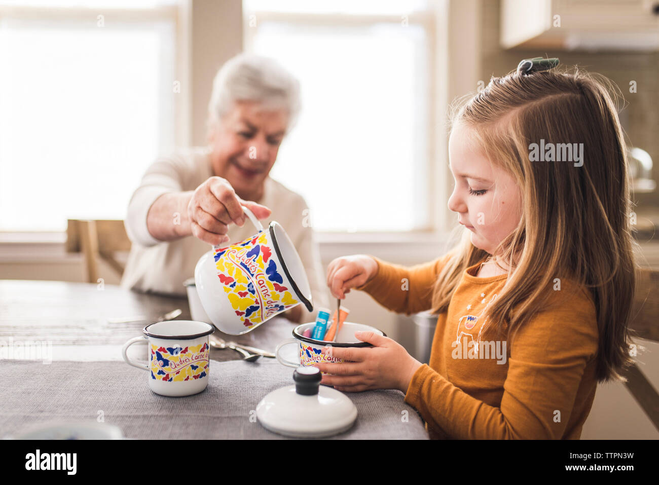 Granddaughter and great grandmother in kitchen playing with tea set Stock Photo