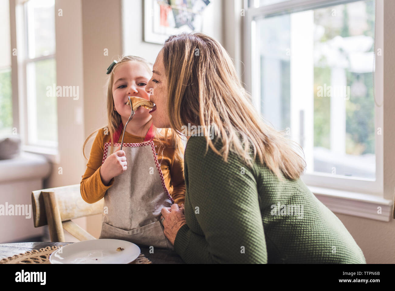 Granddaughter feeding pancakes to grandmother at kitchen table Stock Photo