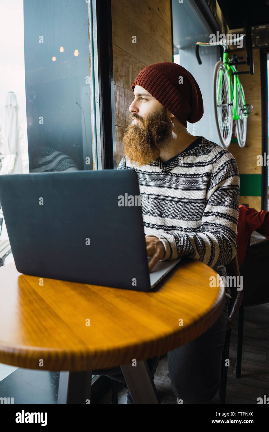 Hipster man looking through window while using laptop computer on table at cafe Stock Photo