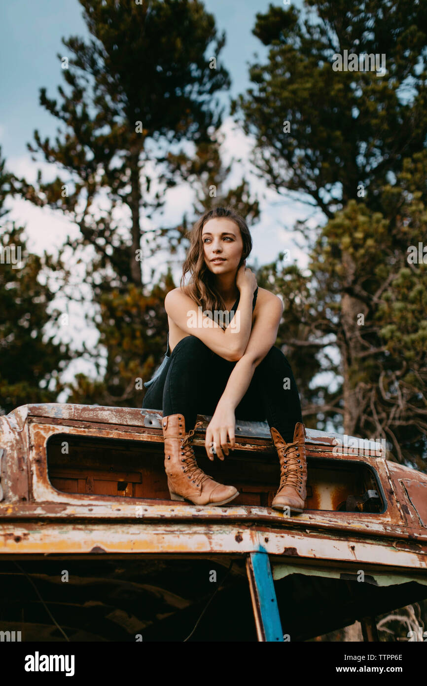 Low angle view of thoughtful young woman looking away while sitting on abandoned vehicle at forest Stock Photo