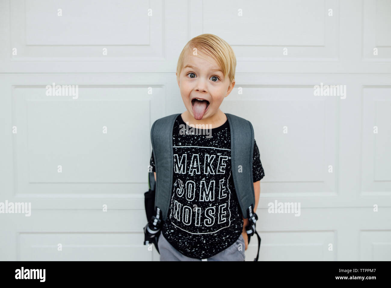 Portrait of boy sticking out tongue while standing with backpack against white wall Stock Photo