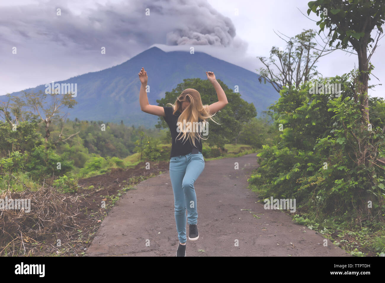 Full length of happy woman dancing on road against smoke erupting volcanic mountain Stock Photo