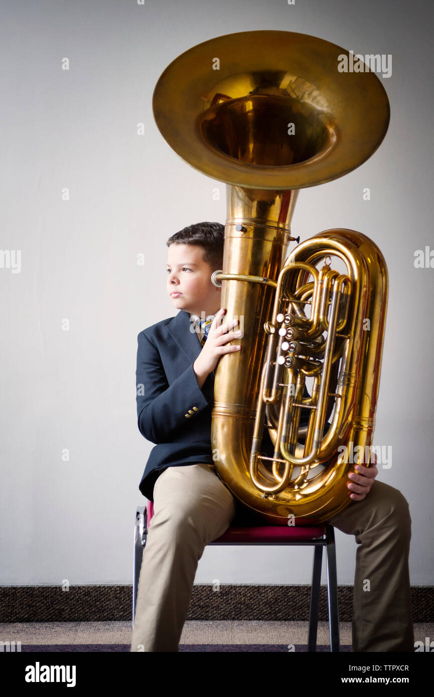 Thoughtful boy holding tuba while sitting on chair against white wall Stock Photo