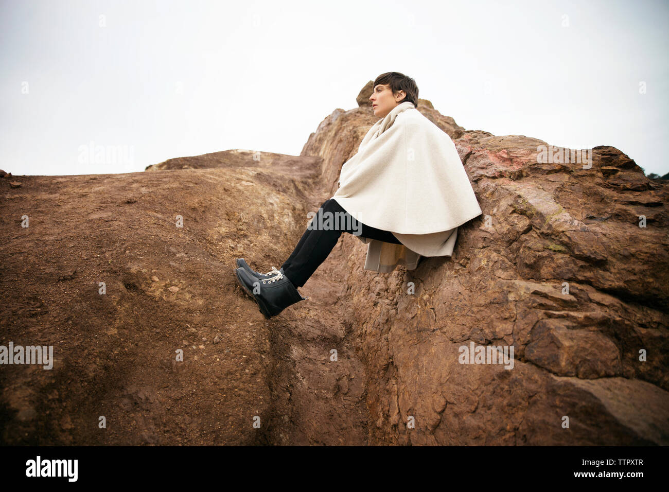 Side view of woman wrapped in jacket sitting on rock against sky Stock ...